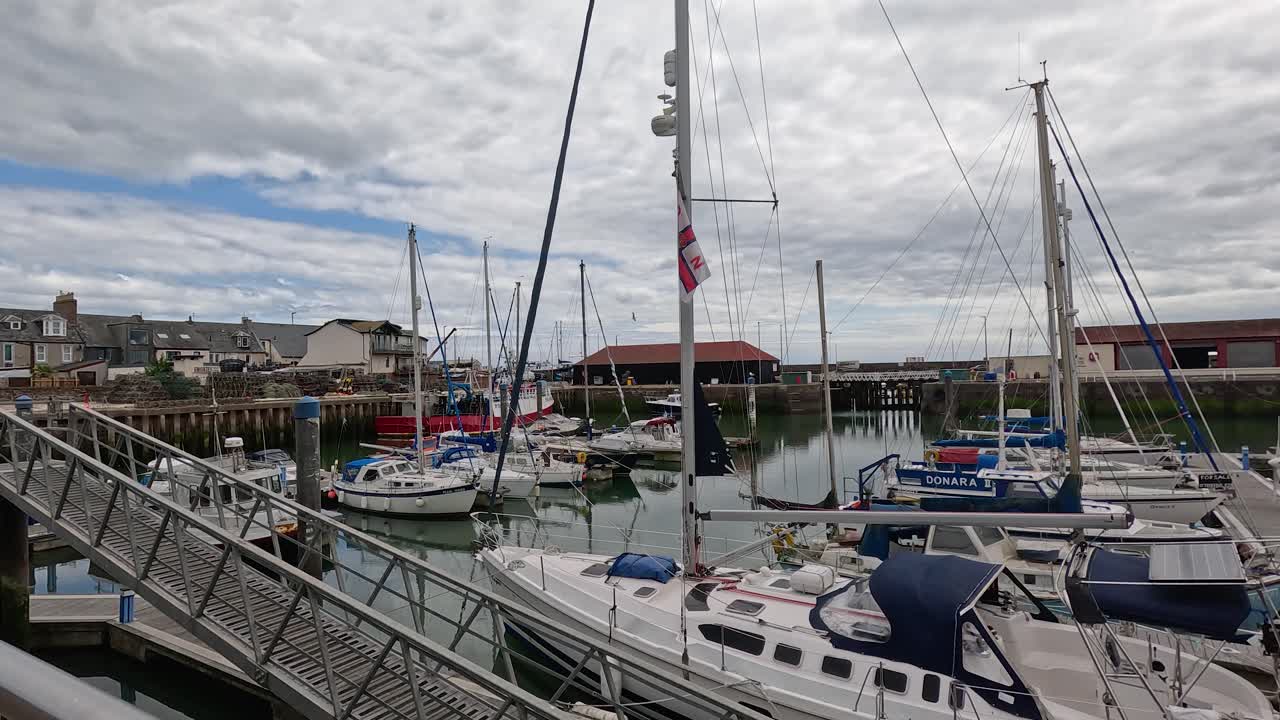 Kamera glir forbi fortøyde seilbåter og yachter i en stille marina under overskyet dagslys