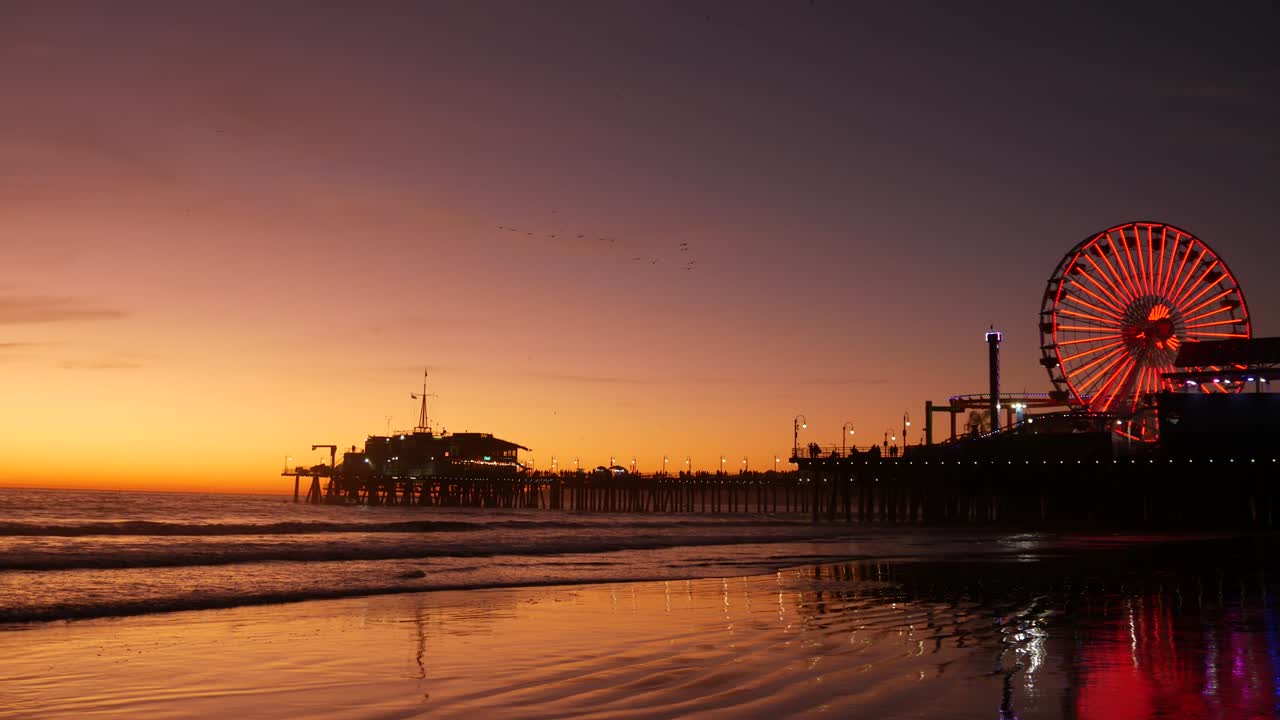 Twilight waves against classic illuminated ferris wheel, amusement park on pier in Santa Monica pacific ocean beach resort. Summertime iconic symbol of California glowing in dusk, Los Angeles, CA USA.
