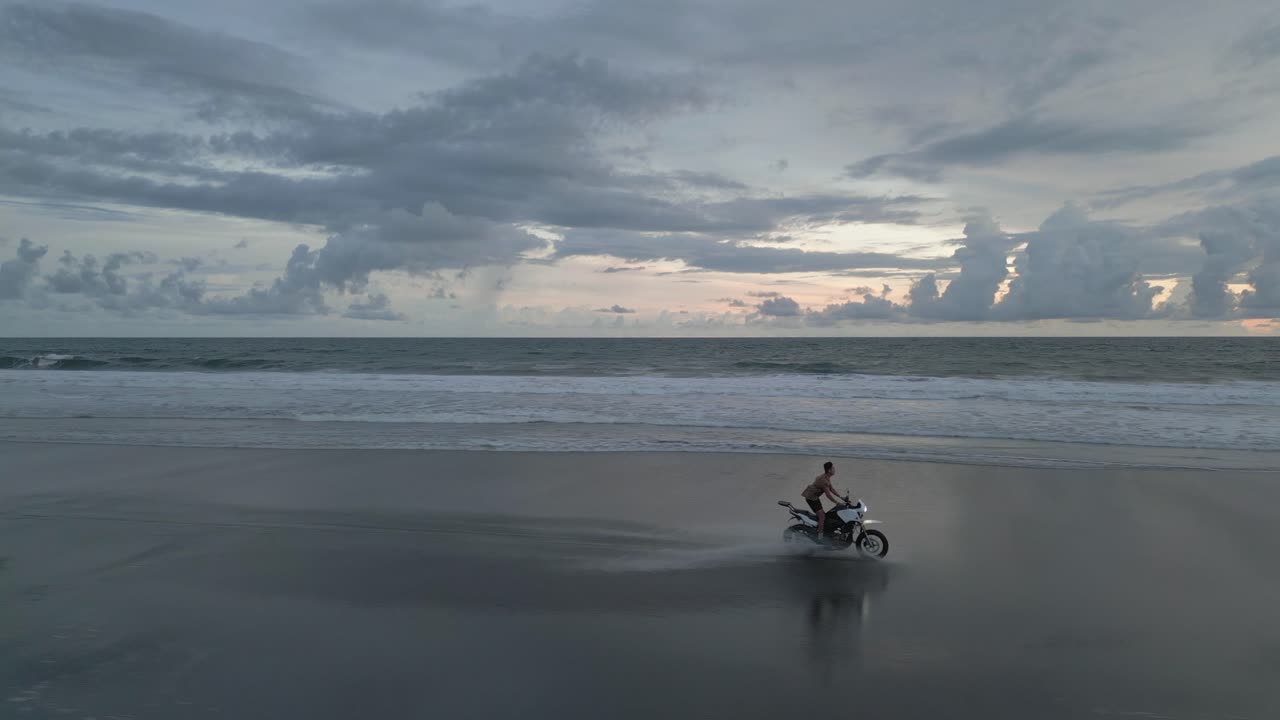 한 남자가 구름진 아침 빛에 모래 바다 해변에서 오토바이를 타고 있습니다.