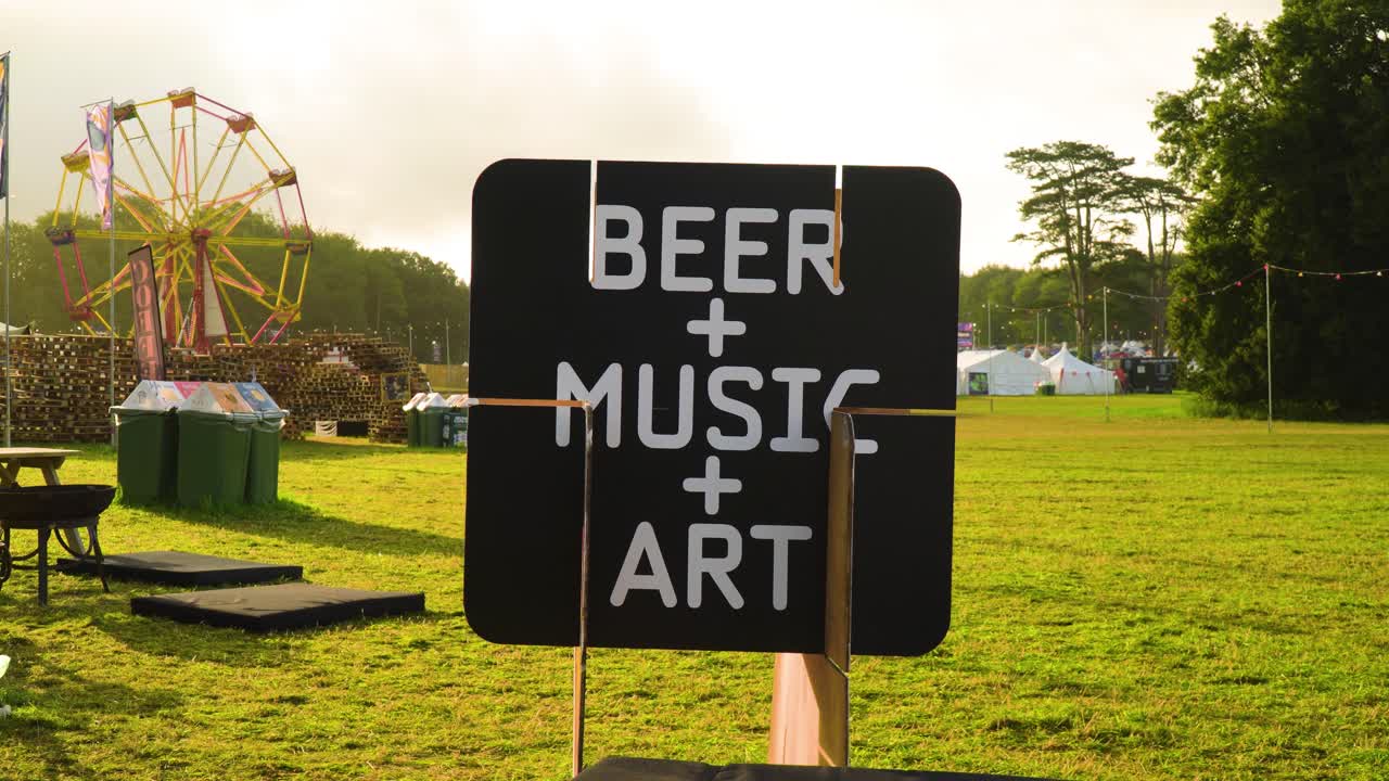 revelando un cartel con música de cerveza y arte escrito en él en un escenario de festival atmósfera mágica con una montaña rusa en el fondo durante la puesta de sol y nadie alrededor en el medio de la naturaleza