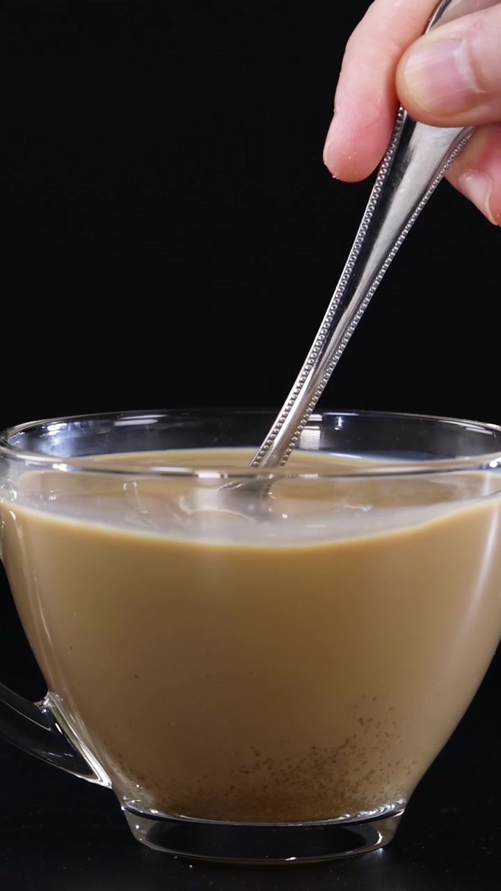 Una mano mescola il caffè con un cucchiaino in una tazza di vetro, su uno sfondo nero, evidenziando il movimento vorticoso.