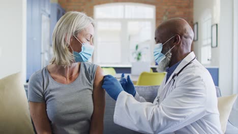 Happy-senior-diverse-woman-and-doctor-wearing-face-masks-in-living-room-sitting-on-sofa,-vaccinating