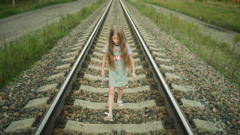young girl with long hair walks on rail track holding grass strand in left hand, nearly tripping on loose gravel between sleepers during quiet stroll in countryside at golden hour