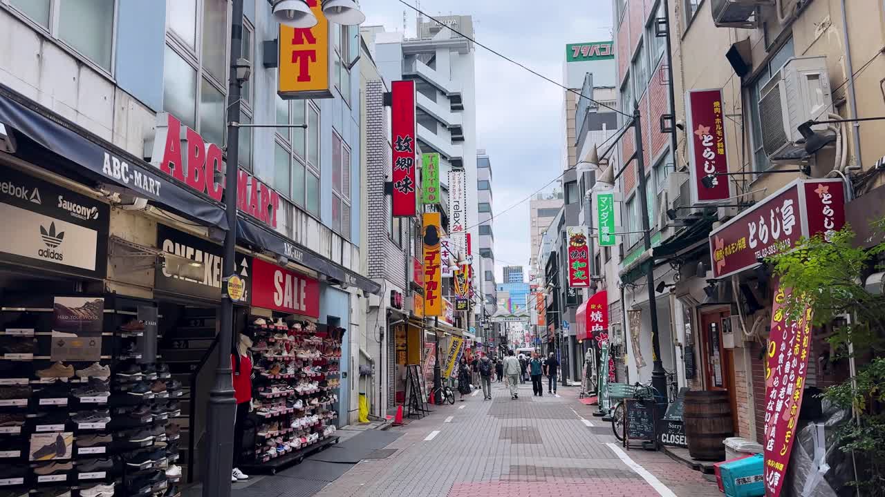 Bustling shopping street with people walking past colorful storefronts on a cloudy day
