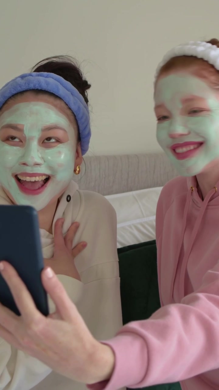 Video verticale: Arriva il telefono, due donne posano per dei selfie sul letto, con maschere verdi e felpe con cappuccio.