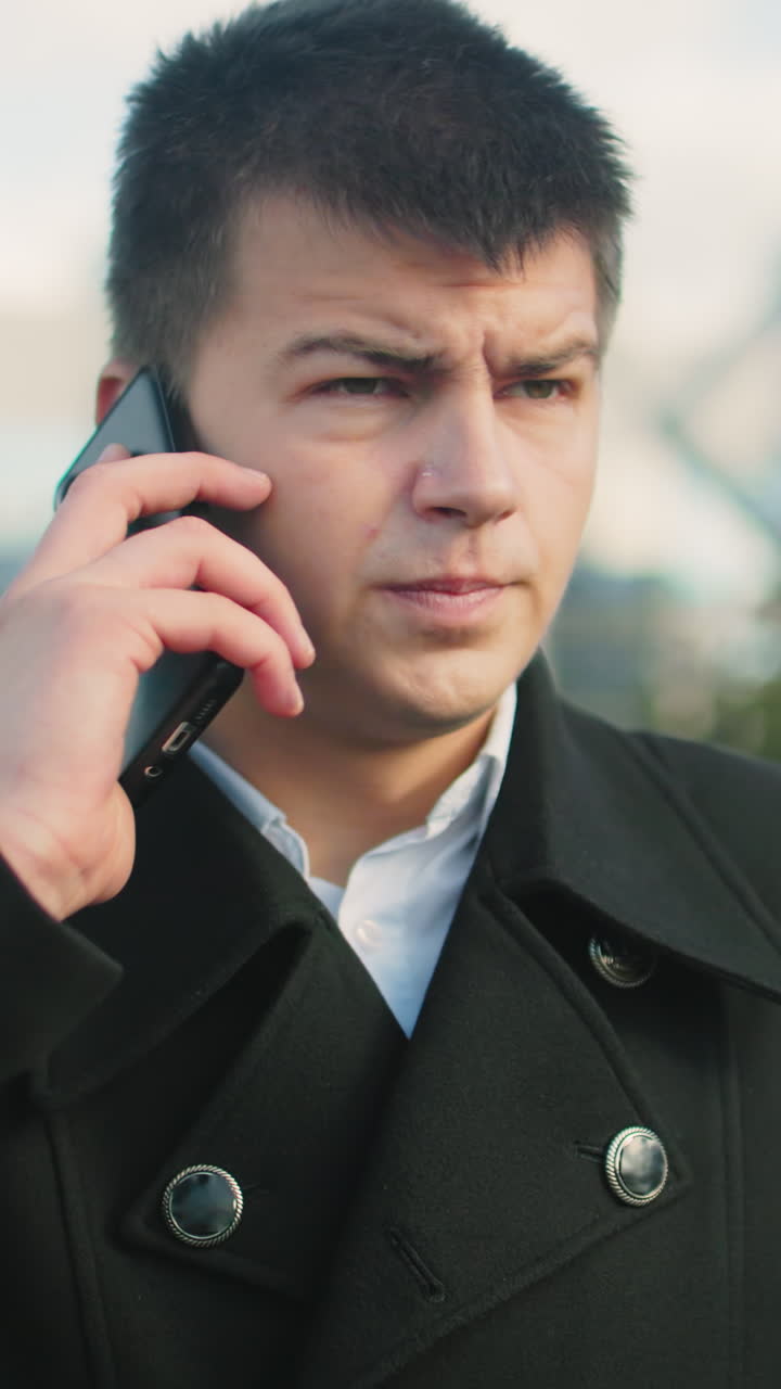 흑색 코트를 입은 남자가 도시 환경에서 밖에서 전화 통화에 응답하는 클로즈업, 배경은 모호한 현대 건물, 초록색, 그리고 지나가는 사람, 남자가 집중하고, 전문적으로 옷을 입은 것처럼 보입니다.