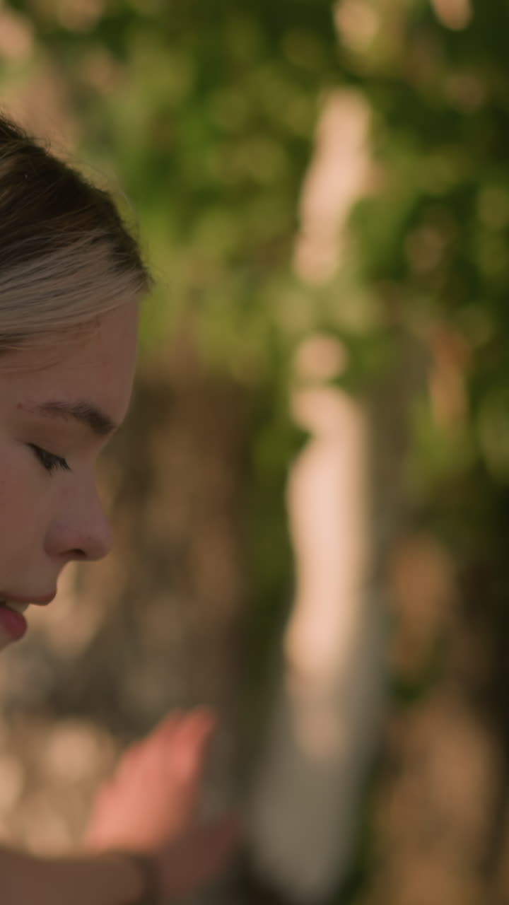 vista lateral de cerca de la joven sonriendo mientras se mueve con gracia como una patinaje como la luz del sol se refleja en ella, el cabello atado hacia atrás con el fondo natural de los árboles