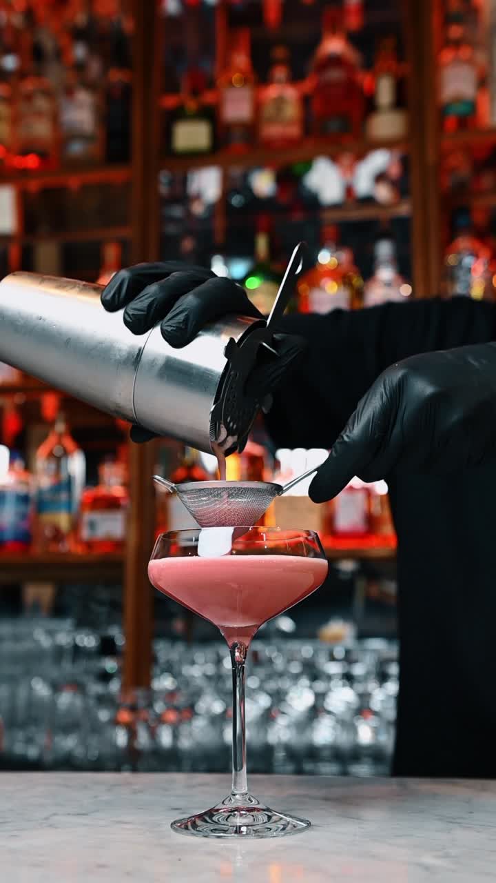 Barman che versa un cocktail rosa attraverso uno strainer in una coppa. Barman professionista con guanti neri che versa una bevanda dallo shaker attraverso un colino in un bicchiere su un bancone del bar in marmo.