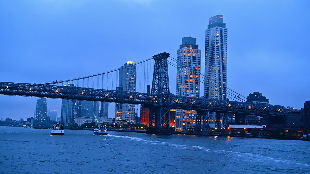 View on the Williamsburg Bridge and the surrounding skyline of New York at sunset time. Two boats go under the bridge. Low angle perspective from the waterscape