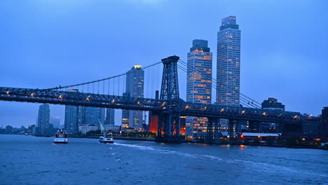 view on the williamsburg bridge and the surrounding skyline of new york at sunset time. two boats go under the bridge. low angle perspective from the waterscape