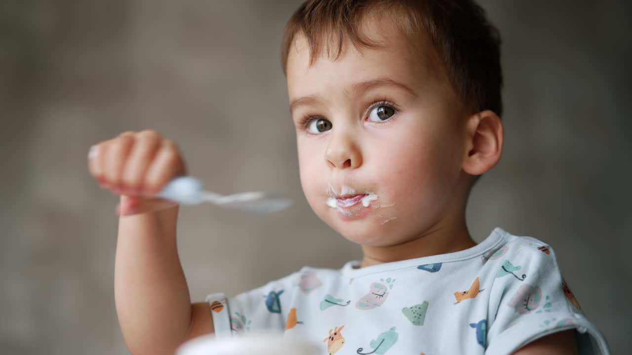 One and a half year old child eating yogurt. Kid finishes the cup and stretches it to camera. Close up. Low angle view.