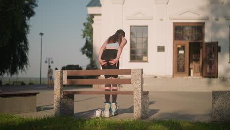 back view of young woman attempting to balance while standing up from bench in roller skates, with white sneakers on ground, urban building in background, and people walking blurred in distance