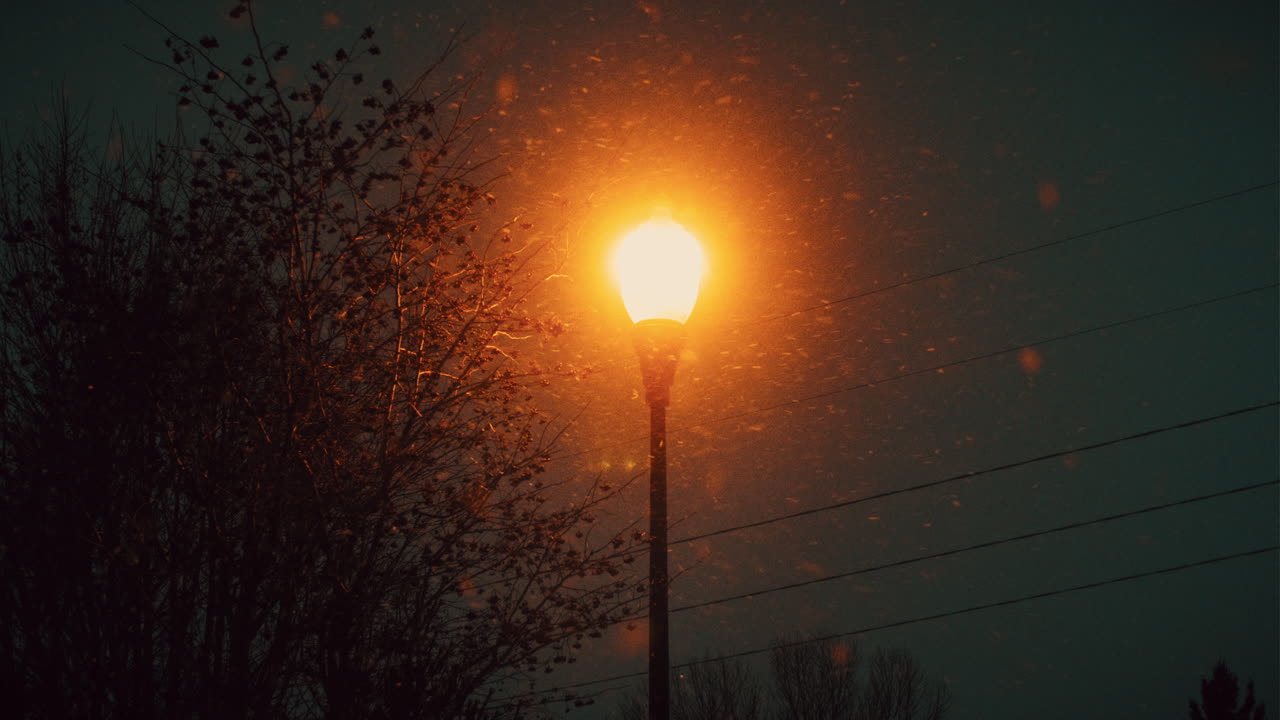 Plano estático de um único poste de luz de rua brilhando em laranja contra o céu noturno enquanto a neve cai durante o inverno no Centro-Oeste