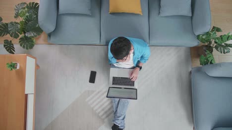 top view of a man holding his chin for thinking and typing on a laptop while sitting on the floor with smartphone in front of a sofa at home