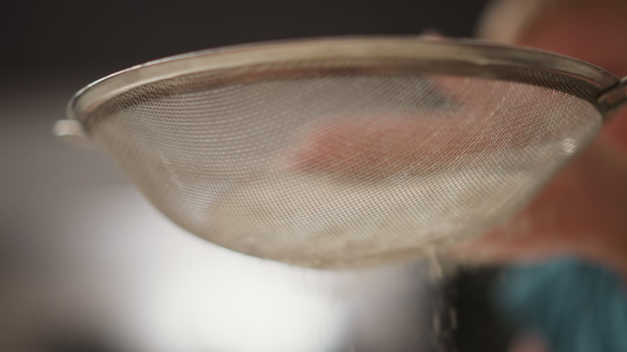 close up hand tapping sieve to sift flour into glass bowl in kitchen setting with soft blurred background showing child silhouette, mixing whisk and rolling pin scattered on counter top