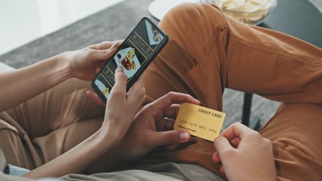 Couple-Ordering-Food-At-Home-Sitting-On-Sofa,-Camera-Focuses-On-Credit-Card-And-Smartphone