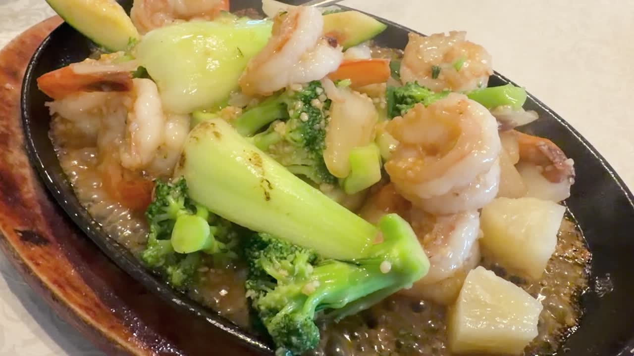 Shrimp, broccoli, bok choy, and vegetables sizzle on a hot plate in bright restaurant lighting