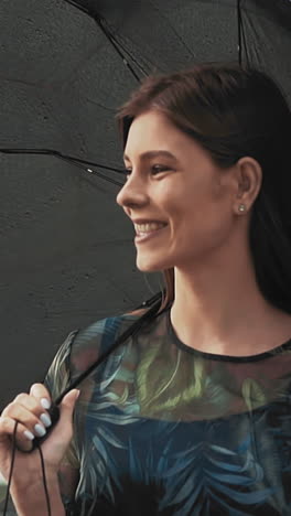 smiling young woman in dress with floral pattern holds black umbrella standing under light rain on city street on warm spring day slow motion closeup