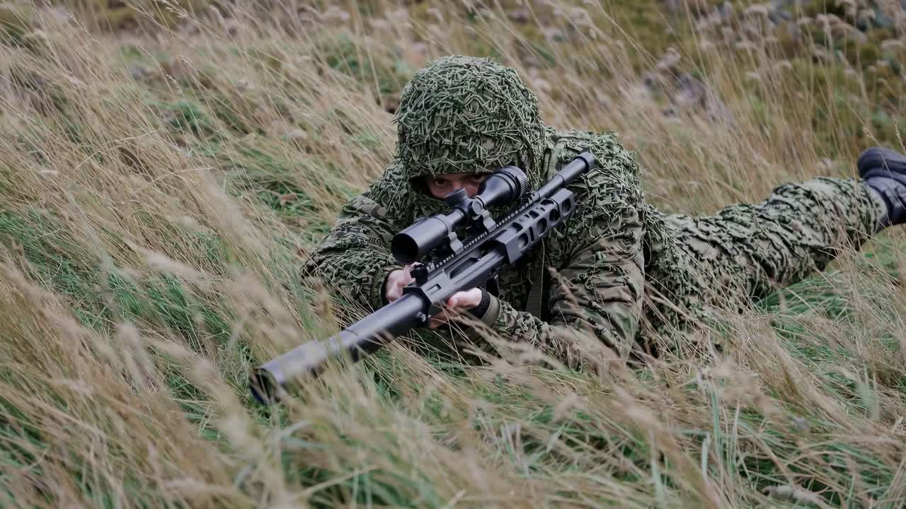 A soldier in camouflage lies prone in tall grass, aiming a sniper rifle