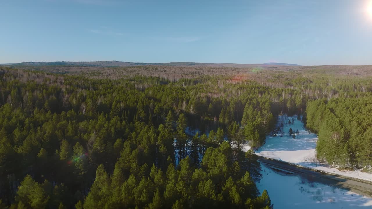 Drönarfilm över vidsträckt barrskog och en frusen flod under låg vintersol, panoramautsikt över vildmarken i Uralbergen, Ryssland, med klar himmel.