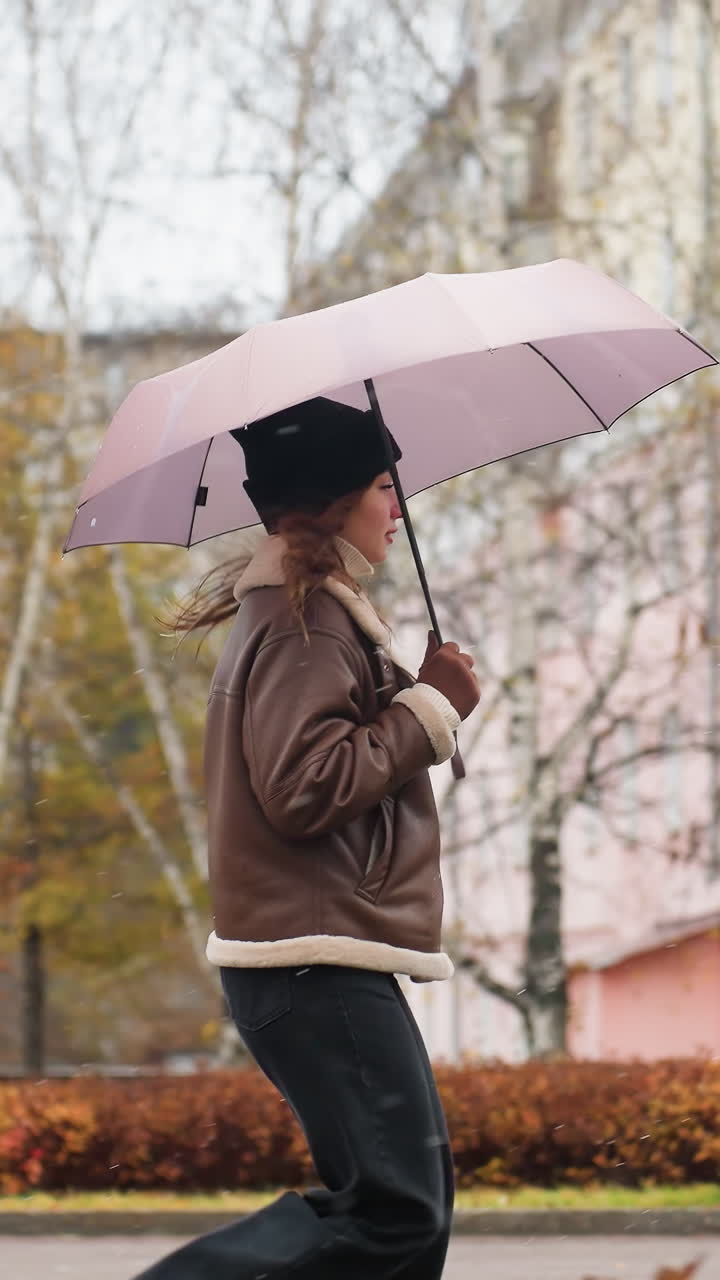 Lady running happily in light snowfall holding umbrella wearing knit cap brown shearling jacket black trousers one hand pocket surrounded by colorful autumn leaves in calm urban setting