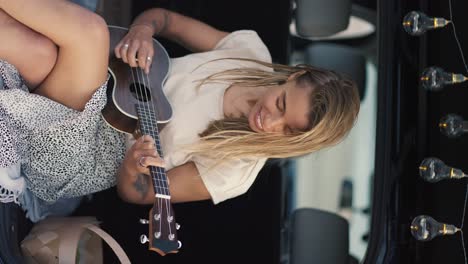 Vertical-video:-A-blonde-girl-plays-the-ukulele-in-the-trunk-of-a-black-car-decorated-with-light-bulbs