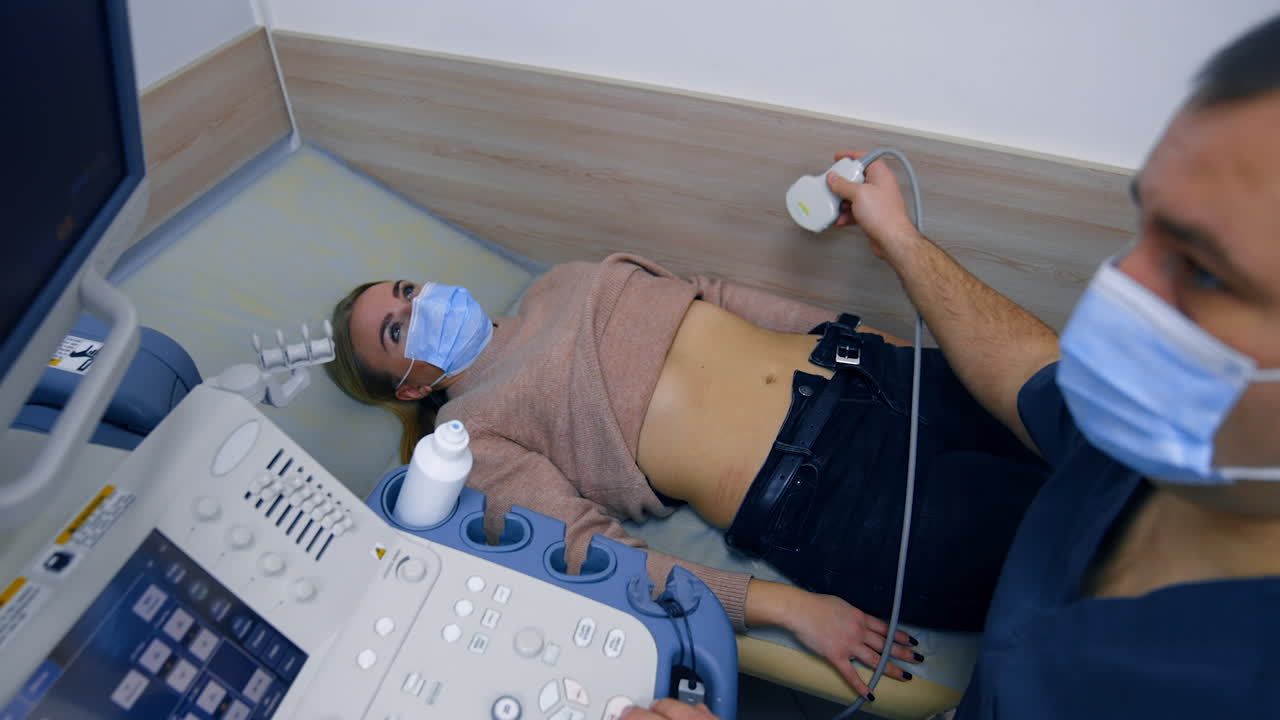 Woman in mask, sweater and jeans lies on the doctor's couch. Male medic puts ultrasonic device on patient's belly side. Top view.