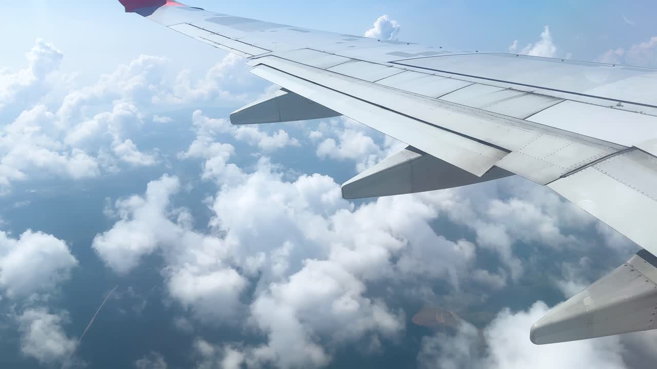 Airplane wing seen from cabin window glides above dense white clouds in bright daylight, with smooth camera movement and clear blue sky