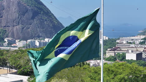 Super-Nahaufnahme-In-Zeitlupe-Der-Brasilianischen-Flagge,-Die-Im-Wind-Weht,-Mit-Den-Liften-Des-Zuckerhuts-Im-Hintergrund