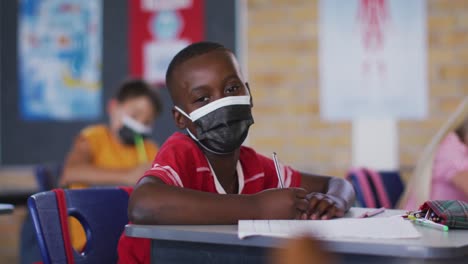 Portrait-of-african-american-schoolboy-wearing-face-mask,-sitting-in-classroom-looking-at-camera