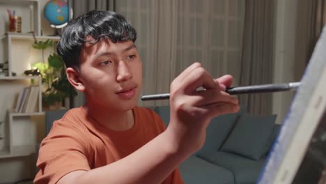 asian artist boy sitting on round wooden chair without backrest holding paintbrush and mixing it with colour before painting on the canvas