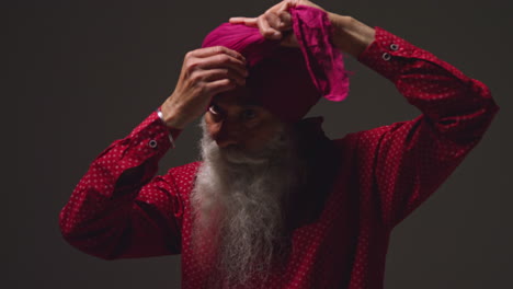 low key studio lighting shot of senior sikh man with beard tying fabric for turban and fastening with pin against dark background