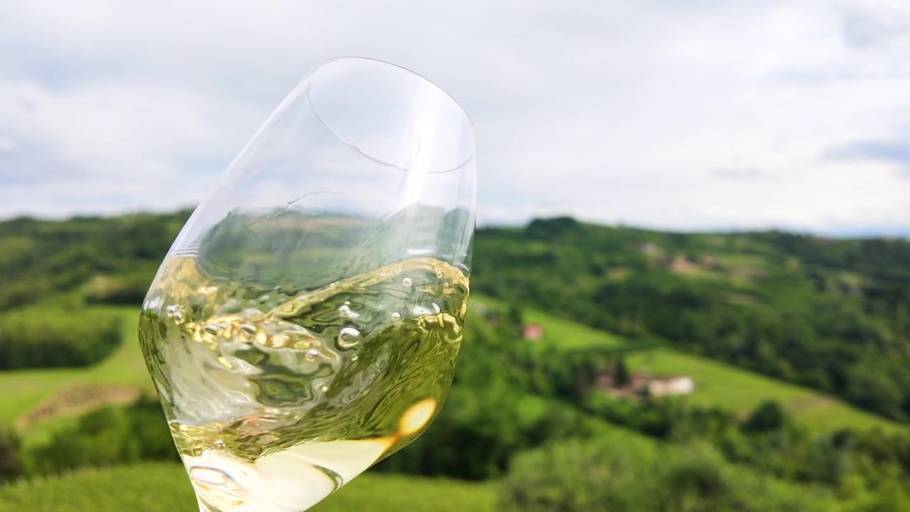Un primo piano di vino bianco che volteggia in un bicchiere sullo sfondo di dolci colline verdi.