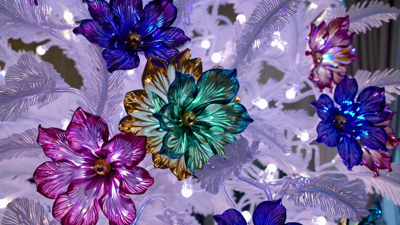 Metallic and colorful flower ornaments adorning white Christmas tree, sparkling with small white lights, embodying festive holiday spirit and seasonal celebration