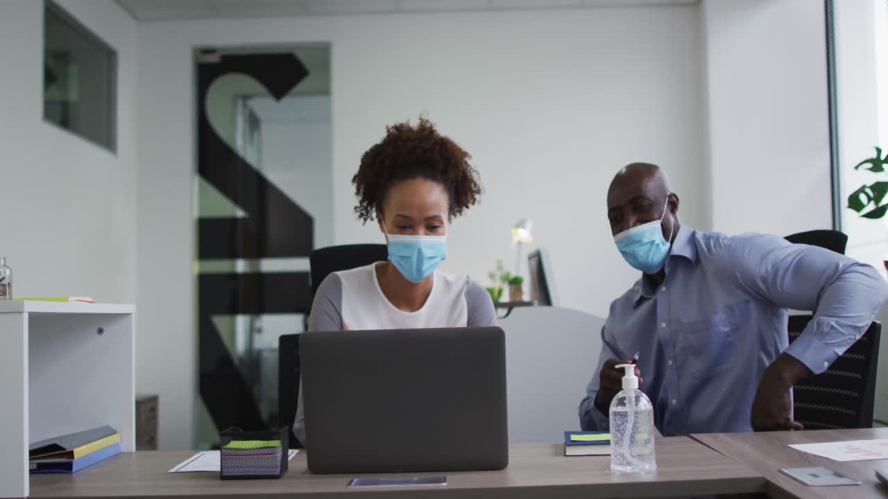 Diverse businessman and businesswoman in face masks discussing, using laptop in office