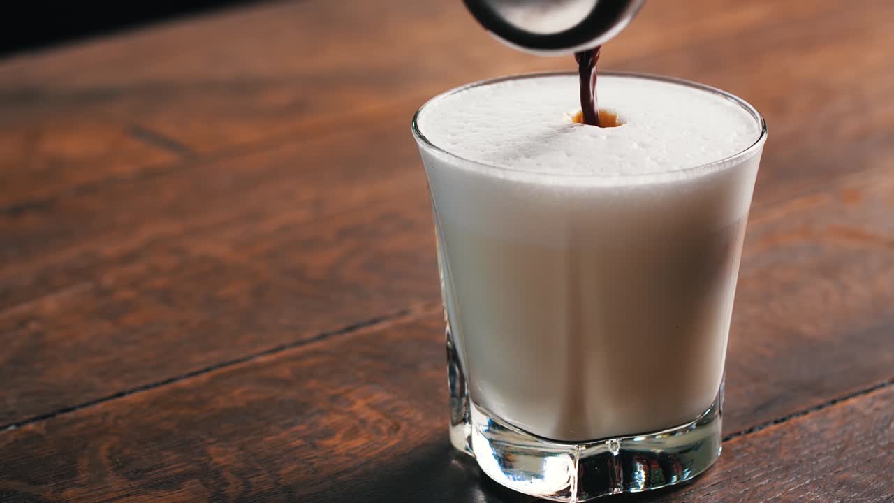 Hot coffee is poured into a glass full of hot milk and foam in slow motion