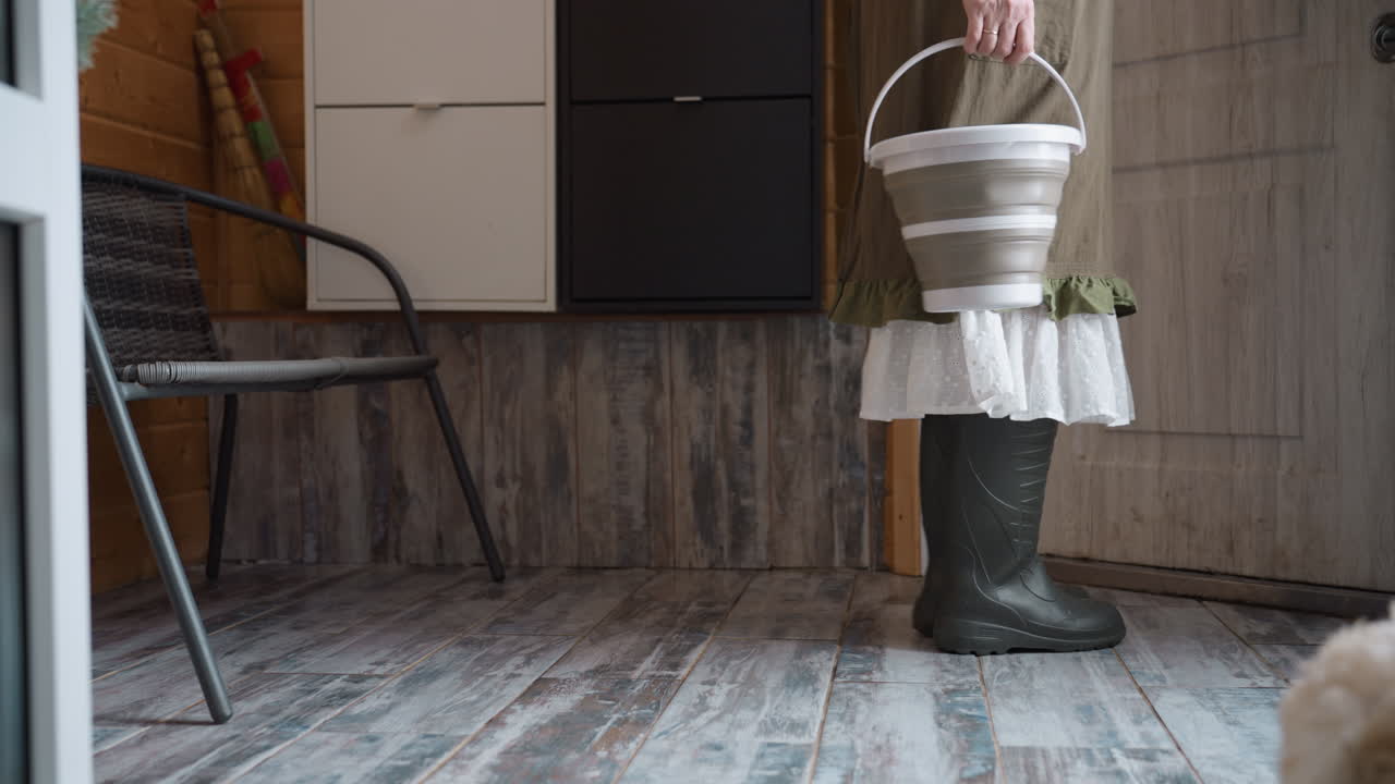 Female gardener standing barefoot on rustic wooden threshold bends down to pick up empty vessel from iron seat beside doorway inside cozy wooden room under warm natural light and textured plank floor