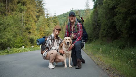 a guy and a girl in hiking clothes, together with their light-colored dog, pose and look at the camera while standing near the road against the backdrop of the forest