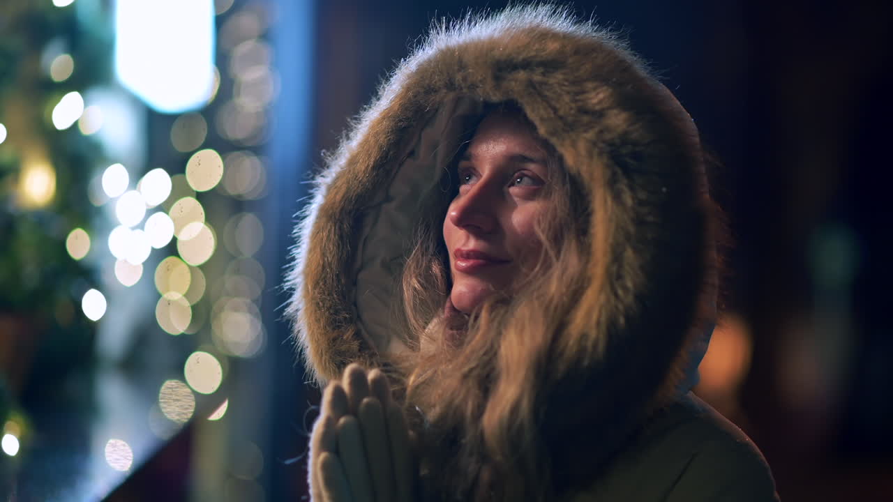 A woman stands in a festive city street, illuminated by holiday lights. She closes her eyes and smiles, embracing the joyful atmosphere while wearing a cozy coat with a fur hood