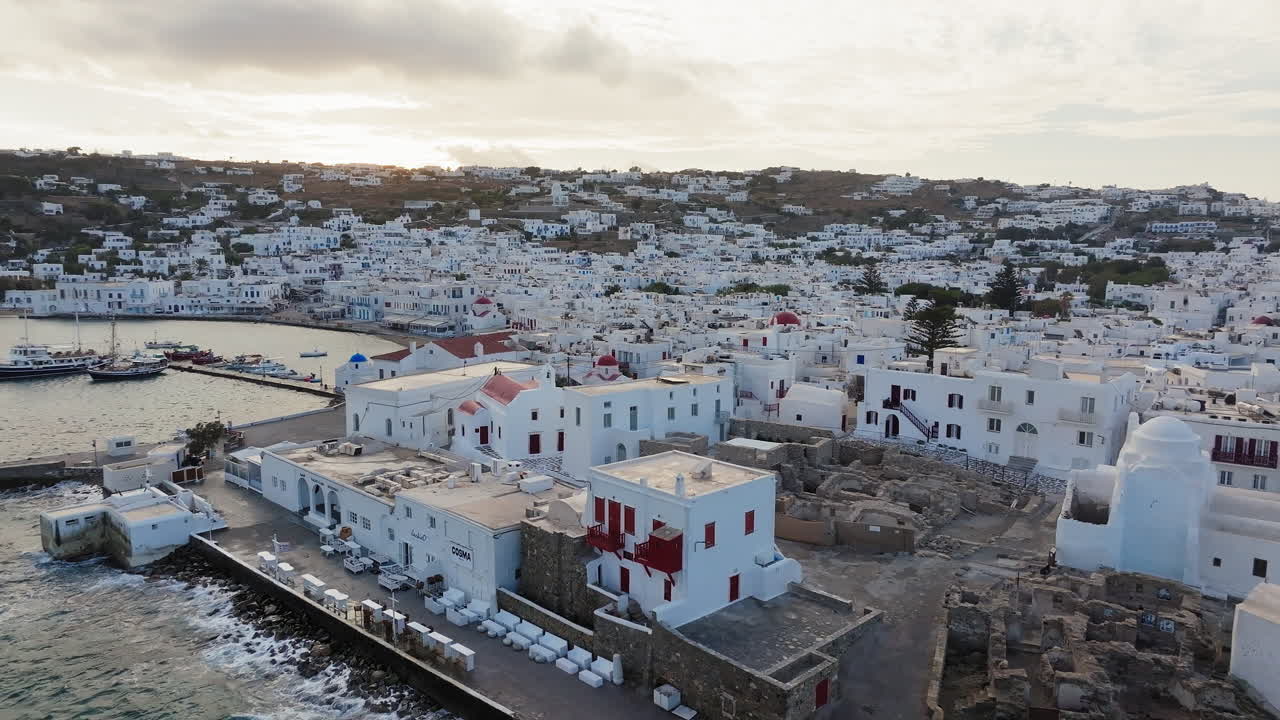 Vista de drone de Chora na ilha de Mykonos ao pôr do sol com casas caiadas de branco e uma cidade turística com porto costeiro