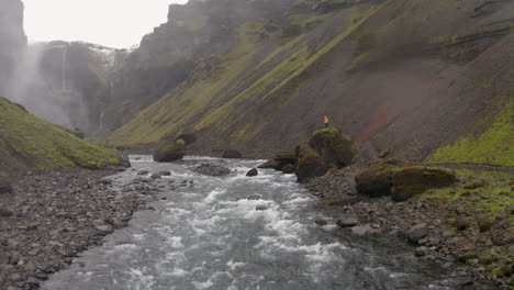 Aerial:-Passing-by-one-person-with-yellow-jacket,-standing-on-a-rock-on-river-bank