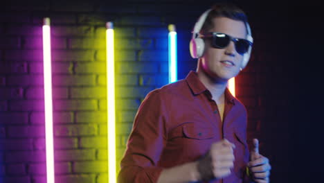 Cheerful-Young-Man-In-Sunglasses-And-White-Headphones-Dancing-And-Listening-To-Music-In-A-Room-With-Colorful-Neon-Lamps-On-The-Wall