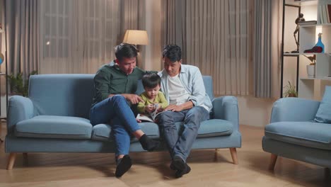 Portrait Of Modern Married Gay Couple Spending Time With Their Son Sitting On Sofa In Living Room Reading Book
