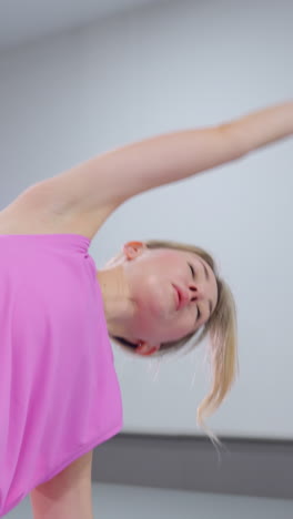 체육관에서 집중적인 스트레칭 운동에 종사하는 여성, 균형을 유지하면서 팔을 펴고, 배경에는 체육 장비와 올바른 자세를 돕는 강사가 있습니다.