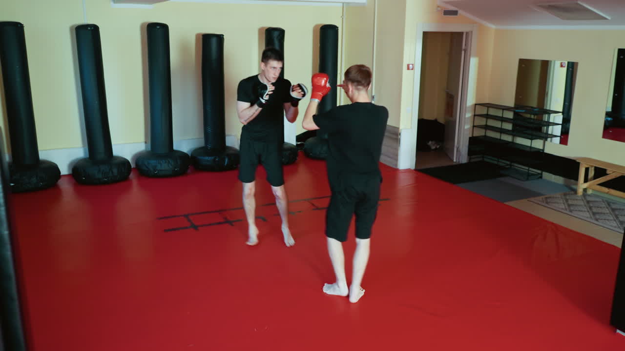 Wrestlers sparring inside gym, wearing gloves and black outfits, practicing combat techniques with raised leg kick and defensive stance on red mat floor surrounded by punching bags during martial training