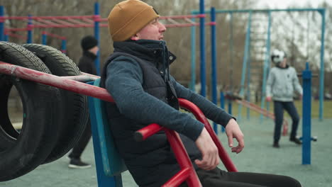 a coach leaning his back on workout equipment, casually shaking his leg, as he looks to watch the two boys playing football in the background