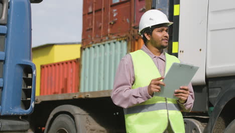 worker wearing vest and safety helmet looking and smiling at camera in a logistics park while holding documents