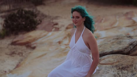 alluring gen z woman in white dress, blue hair, sitting on sandstone rock platform, slomo