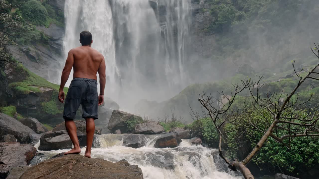 A man stands near the base of Bomburu Ella Waterfalls, surrounded by mist, rocks, and dense greenery in the highlands of Nuwara Eliya, Sri Lanka.