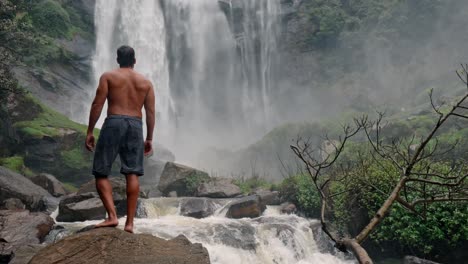 a man stands near the base of bomburu ella waterfalls, surrounded by mist, rocks, and dense greenery in the highlands of nuwara eliya, sri lanka.